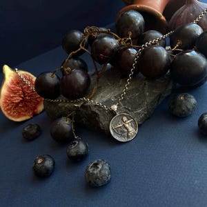 Può includere: Una collana con pendente in argento con un medaglione circolare raffigurante una figura con arco e freccia. La collana è appoggiata su una pietra grigio scuro, circondata da uva scura, mirtilli e un fico tagliato su una superficie blu.