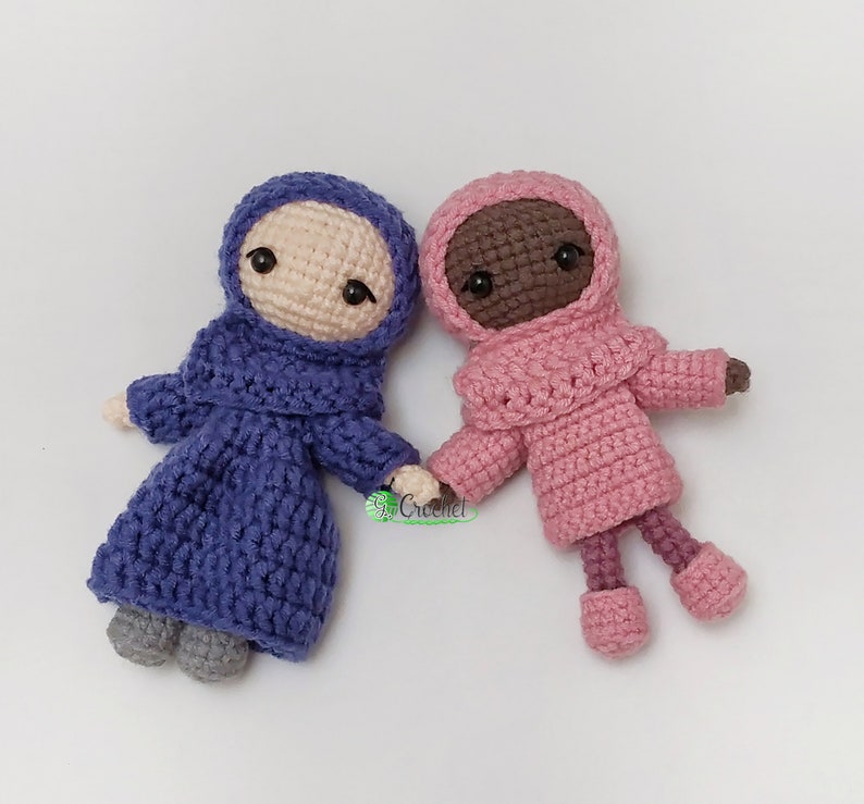 May include: Two crocheted dolls holding hands. One doll is wearing a purple dress and a purple headscarf. The other doll is wearing a pink dress and a pink headscarf. Both dolls have black eyes and brown yarn hair.