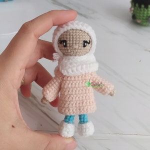 May include: A crocheted doll wearing a white headscarf, pink sweater, blue trousers, and white shoes. The doll has a brown face with black eyes and eyelashes.