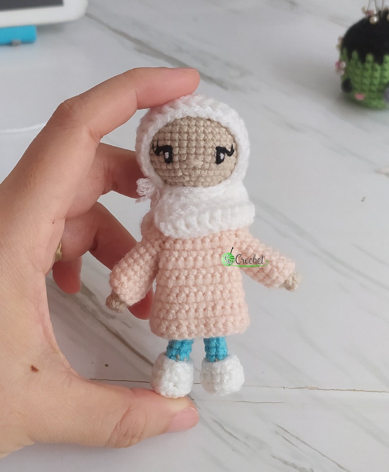 May include: A crocheted doll wearing a white headscarf, pink sweater, blue trousers, and white shoes. The doll has a brown face with black eyes and eyelashes.