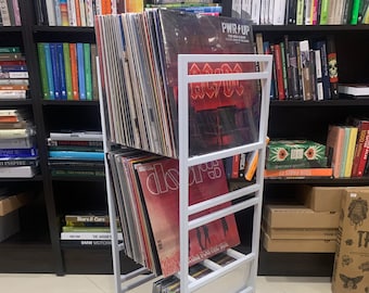 May include: White metal record storage rack with multiple shelves holding vinyl records. The rack is filled with records, including albums by AC/DC, The Doors, and others. The rack is standing on a tiled floor.
