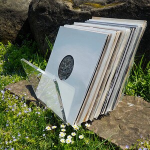 K&ouml;nnte beinhalten: Ein Stapel Schallplatten in einem durchsichtigen Acryl-Schallplattenst&auml;nder. Die Schallplatten sind gr&ouml;&szlig;tenteils wei&szlig; mit einigen schwarzen und braunen Akzenten. Der St&auml;nder steht auf einem Stein.