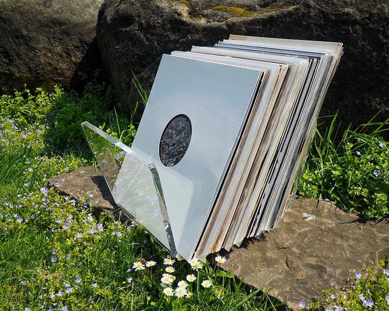 K&ouml;nnte beinhalten: Ein Stapel Schallplatten in einem durchsichtigen Acryl-Schallplattenst&auml;nder. Die Schallplatten sind gr&ouml;&szlig;tenteils wei&szlig; mit einigen schwarzen und braunen Akzenten. Der St&auml;nder steht auf einem Stein.