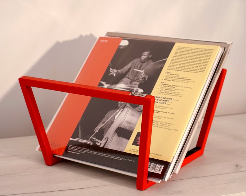 K&ouml;nnte beinhalten: Ein roter Metall-Plattenst&auml;nder mit einem Stapel Schallplatten. Die oberste Platte zeigt ein Schwarzwei&szlig;foto eines Mannes, der Bass spielt. Das Plattencover hat den Text "Johnny Hodges Album with Jon Jones".