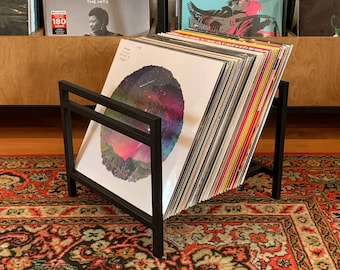 May include: Black metal record stand with a stack of vinyl records. The stand has a rectangular shape and is made of metal. The records are stacked vertically and are of various colors and sizes. The stand is on a red and black patterned rug.