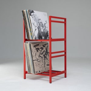 May include: Red metal record rack with four shelves holding vinyl records. The records have black and white illustrations and text.