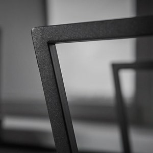 May include: Close-up of a section of a black metal chair frame. The frame has a textured surface and a geometric design. The chair appears to be modern and minimalist in style. The background is blurred, focusing attention on the chair's details.