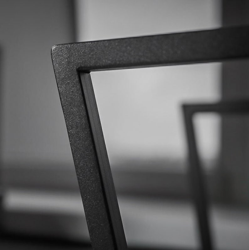 May include: Close-up of a section of a black metal chair frame. The frame has a textured surface and a geometric design. The chair appears to be modern and minimalist in style. The background is blurred, focusing attention on the chair's details.