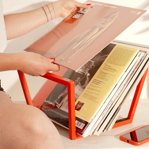 K&ouml;nnte beinhalten: Ein leuchtend orangefarbener Schallplattenst&auml;nder h&auml;lt einen Stapel Schallplatten. Der St&auml;nder ist aus Metall und hat ein schlichtes, modernes Design. Eine Person h&auml;lt eine Schallplatte in der Hand.