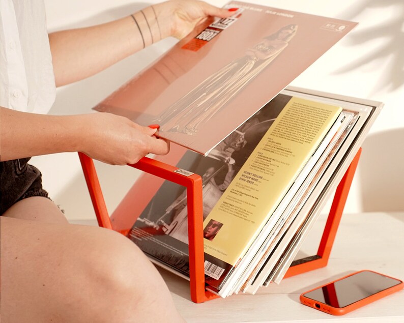 K&ouml;nnte beinhalten: Ein leuchtend orangefarbener Schallplattenst&auml;nder h&auml;lt einen Stapel Schallplatten. Der St&auml;nder ist aus Metall und hat ein schlichtes, modernes Design. Eine Person h&auml;lt eine Schallplatte in der Hand.