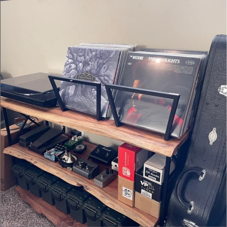 May include: A wooden audio equipment shelf with a live edge design. It holds vinyl records, a turntable, and various guitar effects pedals. The records are displayed in black metal stands. A guitar case is on the right.