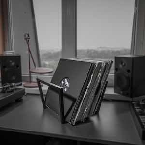 May include: A black metal record holder displaying a stack of vinyl records. The holder has a modern, angular design and sits on a dark surface. Two black speakers and a turntable are visible in the background, suggesting a music listening setup.