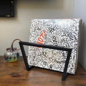 May include: A black metal record stand holding a vinyl record album. The album cover is white with the word "RIOT!" in black and red, repeated across the surface. The album is titled "Paramore".