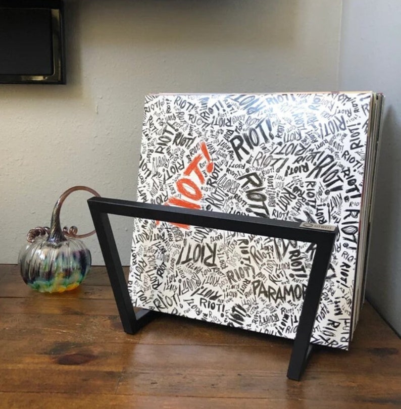 May include: A black metal record stand holding a vinyl record album. The album cover is white with the word "RIOT!" in black and red, repeated across the surface. The album is titled "Paramore".