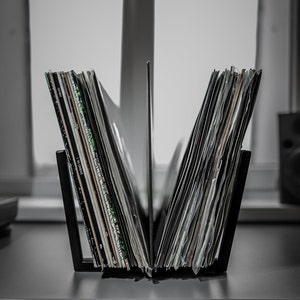 May include: A black metal record holder filled with vinyl records. The holder is divided into two sections, each holding a stack of records. The record sleeves display various colors and designs, with some text visible. The holder sits on a dark surface.