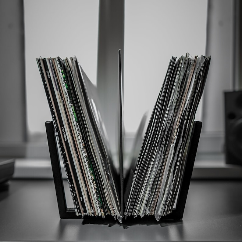 May include: A black metal record holder filled with vinyl records. The holder is divided into two sections, each holding a stack of records. The record sleeves display various colors and designs, with some text visible. The holder sits on a dark surface.