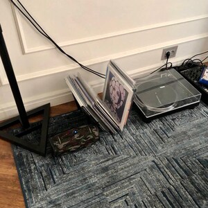 May include: A black turntable with a clear cover sits on a blue patterned rug. Vinyl records are propped up next to a camouflage-patterned speaker. A black stand with a triangular base is in the foreground.