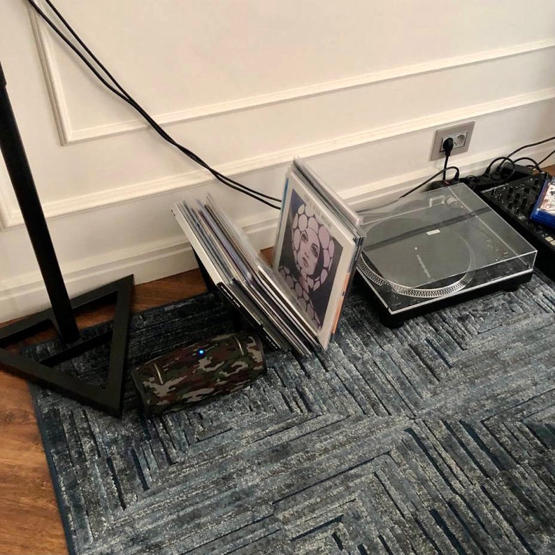 May include: A black turntable with a clear cover sits on a blue patterned rug. Vinyl records are propped up next to a camouflage-patterned speaker. A black stand with a triangular base is in the foreground.