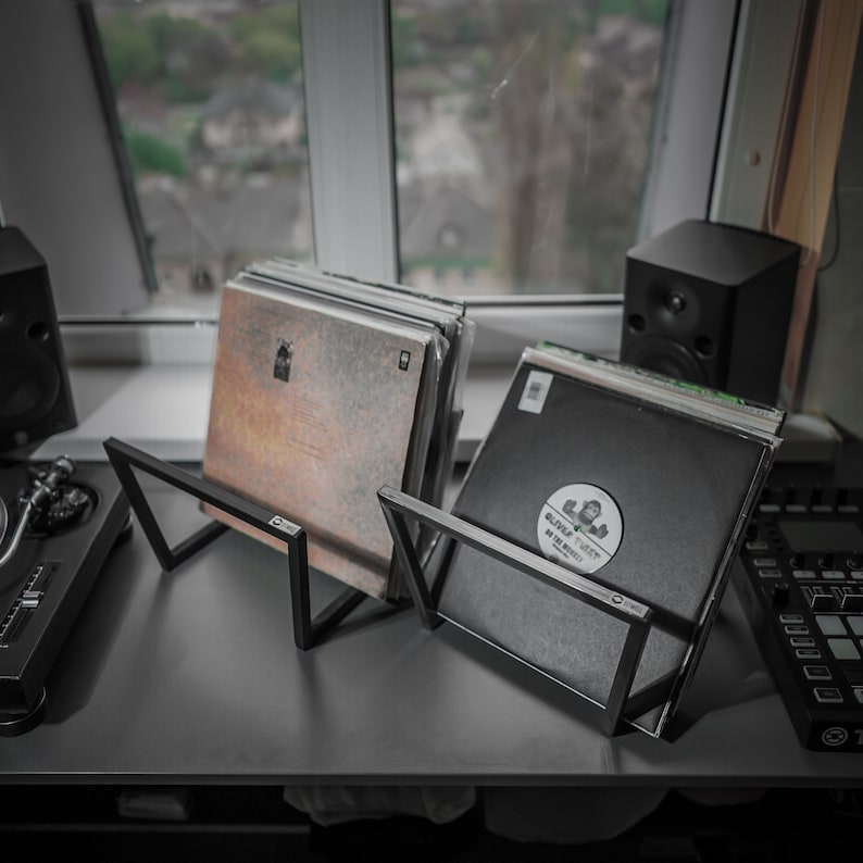 May include: Two black metal record holders displaying vinyl records. The holders have a minimalist, angular design. One record has a circular sticker with text. The holders are on a gray surface near a window.