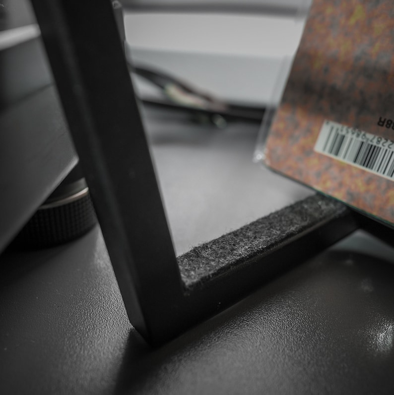 May include: Close-up of a black metal frame with a gray felt pad on the bottom edge. A partially visible object with a barcode is in the background. The frame is on a dark gray surface.