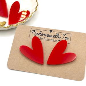 Peut inclure: Deux boucles d'oreilles rouges en forme de cœur sur une carte marron avec le texte "Mademoiselle No". Les boucles d'oreilles sont présentées sur une surface blanche avec une assiette décorative dorée.