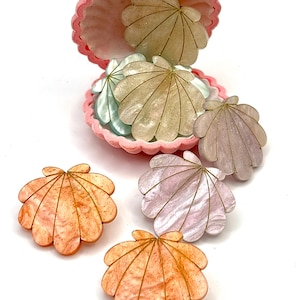 Peut inclure: Un récipient rose en forme de palourde contenant plusieurs ornements irisés en forme de coquillage dans des tons pêche, lavande et crème. Chaque coquillage présente une finition pailletée subtile et de fines lignes dorées partant de la base.