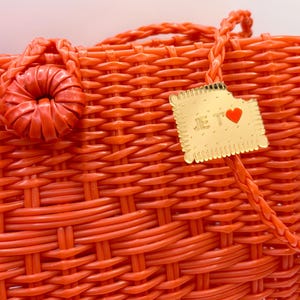 Peut inclure: Gros plan sur un sac à main orange tissé avec une poignée tressée et une fleur décorative. Une étiquette dorée avec les mots "JE T'" et un cœur rouge est attachée au sac.