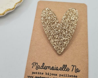 Puede incluir: Un broche dorado en forma de corazón con purpurina sobre una tarjeta marrón con el texto "Mademoiselle No". El broche es un accesorio brillante. Un plato de cerámica blanco con ribete dorado está en el fondo.