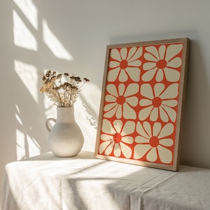 May include: A white ceramic vase with dried flowers sits on a white linen tablecloth next to a framed print of a red and white floral pattern.
