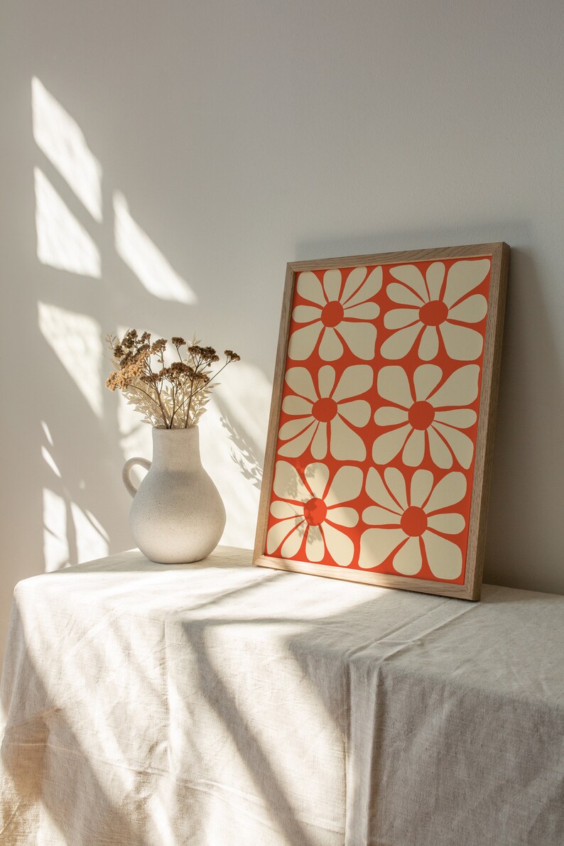 May include: A white ceramic vase with dried flowers sits on a white linen tablecloth next to a framed print of a red and white floral pattern.