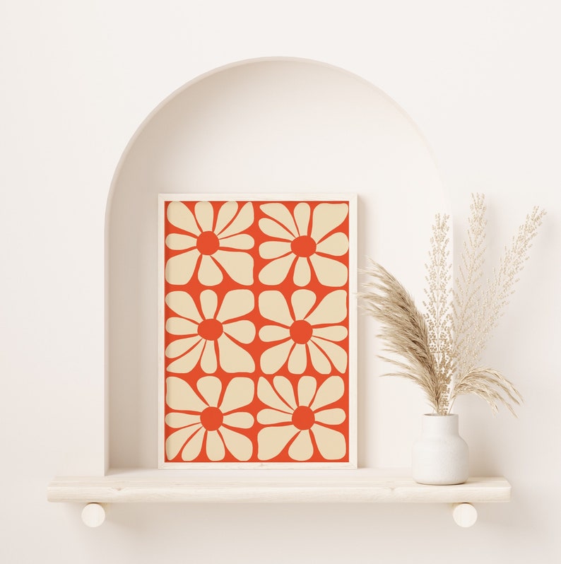May include: A framed print with a geometric pattern of white flowers on a red background. The print is displayed on a white shelf with a vase of dried pampas grass.