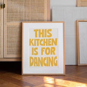 Puede incluir: Una impresión enmarcada con el texto "This kitchen is for dancing" en letras amarillas sobre un fondo blanco.