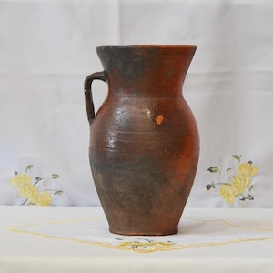 May include: A rustic, brown ceramic jug with a handle. The jug has a wide base that tapers to a narrower neck and flared rim. The surface has a mottled appearance with shades of brown and orange, suggesting it is handmade.
