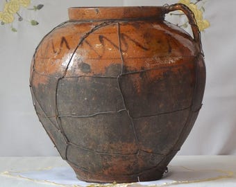 May include: A large, antique ceramic jar with a handle, featuring a two-tone color scheme of brown and dark brown. The jar is encased in a wire mesh, and has decorative markings near the top. The jar is likely used for storage.