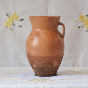 May include: A brown clay pitcher with a handle. The pitcher has a slightly darker brown color at the bottom.