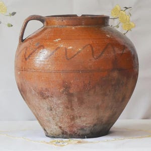 May include: A large, antique, brown ceramic jug with a handle. The jug has a wide body and a narrow neck with a small opening. A wavy, dark brown line decorates the upper portion of the jug. The jug has a rustic, aged appearance.