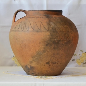 May include: A large, round, terracotta-colored ceramic jug with a handle. The jug features a dark rim and a decorative band of black lines around the upper portion. The surface has dark patches, suggesting age and use. A rustic, handmade pottery piece.