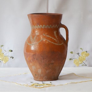 May include: A brown clay pitcher with a white design. The pitcher has a rounded body and a handle.