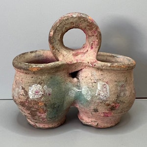 May include: A double-handled ceramic pot with a handle in the shape of a ring. The pot is painted in shades of pink, brown, and green with white accents.