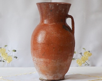 Op de afbeelding: Een terracotta kruik met een enkele handgreep. De kruik is roodbruin van kleur en heeft een licht onregelmatig oppervlak.