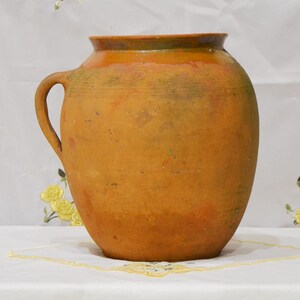 May include: A large, brown, earthenware jug with a wide mouth and a single handle. The jug has a rustic, handmade appearance and is decorated with a simple, incised design around the rim.