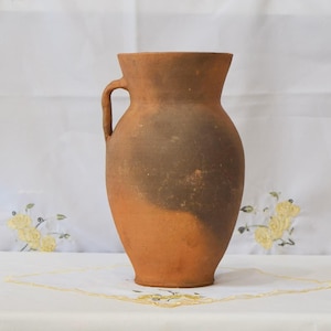 May include: A rustic, earthenware jug with a handle. The jug has a rounded body and a narrow neck. The color is a mix of brown and orange, with a darker band around the middle. The jug is likely handmade and has a vintage aesthetic.