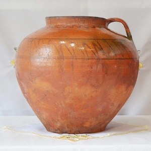 May include: A large, brown clay pot with a wide mouth and a single handle. The pot has a rustic, hand-crafted look and is decorated with a simple, hand-painted design.