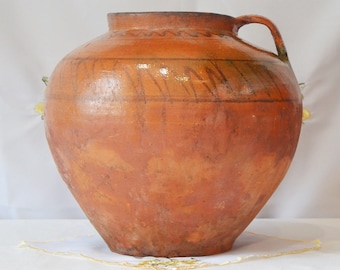 Op de afbeelding: Een grote, bruine kleipot met een brede opening en een enkele handgreep. De pot heeft een rustieke, handgemaakte uitstraling en is versierd met een eenvoudig, handgeschilderd ontwerp.