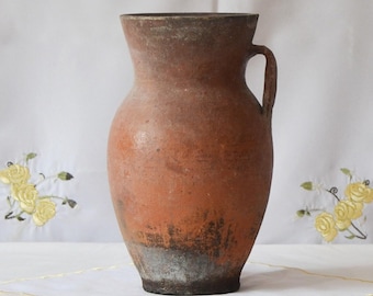 May include: A terracotta vase with a handle, showing signs of wear and age. The vase is a light brown color with a darker brown rim and base.