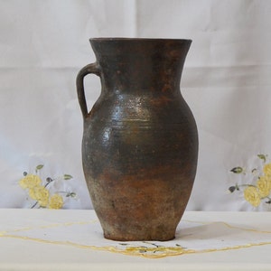 Könnte beinhalten: Ein brauner Tonkrug mit Henkel. Der Krug hat ein rustikales, gealtertes Aussehen.
