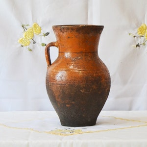 May include: A brown clay pitcher with a handle. The pitcher has a dark brown glaze on the bottom half and a lighter brown glaze on the top half.