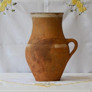 Op de afbeelding: Een bruine aardewerken kruik met een handvat. De kruik heeft een brede basis die smaller wordt naar een hals, met een witte band rond het midden en de rand. De kruik is waarschijnlijk handgemaakt en heeft een vintage uitstraling.