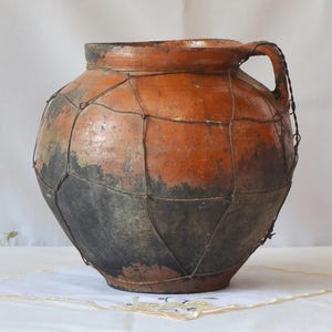 May include: A rustic, antique terracotta jar with a handle, featuring a wire mesh overlay. The jar has a rounded shape, with a dark, weathered appearance on the lower half and a lighter, orange-brown hue on the upper portion.