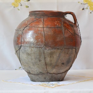 May include: A large, round, terracotta pot with a wire mesh covering. The pot has a brown and gray color with a worn, rustic appearance.
